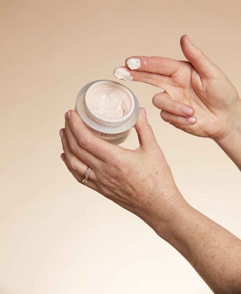 Open jar of Erasa SPF-30 Lifting/Sculpting Créme. A hand holds the jar with one hand and the other hand the product on their fingertips displaying the texture consistency  of the créme.